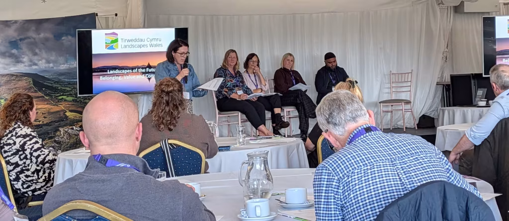 Panel discussion about "Landscapes of the Future: Belonging, Value and Connection" with a speaker at a microphone and four seated panelists, with an audience in the foreground.